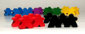 In the foreground is a group of four red meeples next to a group of four black meeples. In the background can be seen a cluster of meeples, with the ones on the left in blue, the ones next to those in purple, then green, and the ones on the right are yellow.