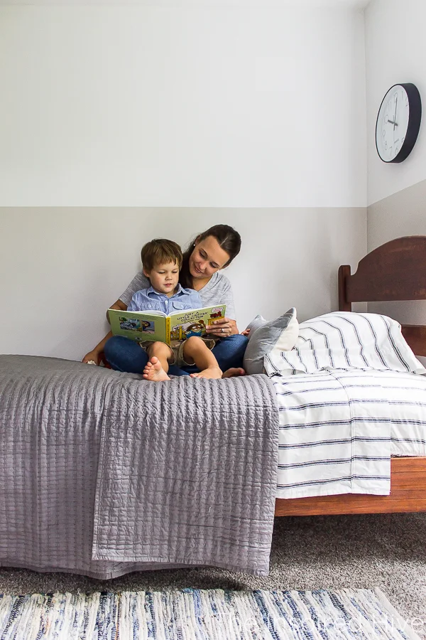 Reading books to toddler at bedtime. Boy's bedroom with antique bed, grey quilt, denim rug. 