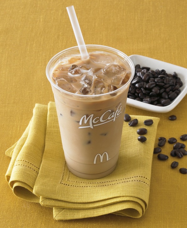 iced coffee in McDonald’s cup with straw on folded gold napkin with coffee beans in small white dish and more strewn artfully about the table