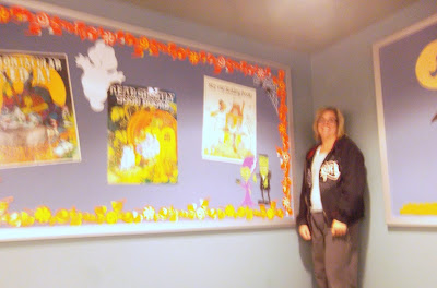 Woman stands to the right of a bulletin board decorated with Halloween posters, die-cut characters and pumpkin shape borders.