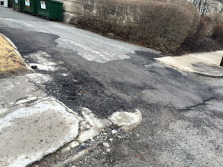 Photograph. Diagonal view of a roughly patched street and cracked sidewalks.