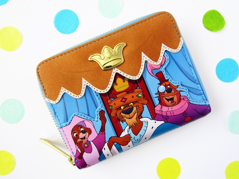 A photo of a blue and yellow Loungefly wallet with characters from Disney's Robin Hood printed on the back photographed from above on a white background surrounded by confetti. The back of the wallet features images of Maid Marian (a vixen in pink and white clothing), Prince John the lion, and Little John the brown bear dressed as Sir Reginald, Duke of Chutney, sitting in the royal box watching an archery tournament.