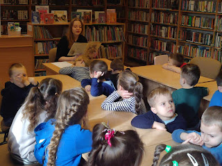 Wnętrze biblioteki. Na pierwszym planie dwa podłużne stoły z jasnego drewna, przy których siedzi grupa przedszkolaków. Dzieci patrzą w stronę bibliotekarki i  słuchają bajki o Kocie w Butach. W tle widać bibliotekarkę, która siedzi i czyta im książkę. Za nią znajdują się regały pełne książek i wystawka na temat kotów. Na zielonych ścianach można dostrzec tablicę korkową i plakat promujący czytelnictwo. Obok tablicy stoi komputer.