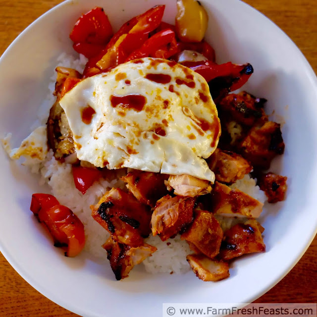 a square image of a fried egg atop grilled chicken, eggplant, and peppers over rice