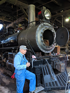 A docent in denim coveralls with a red bandana and a railway cap stands with his foot on the rail as he talks about this engine.