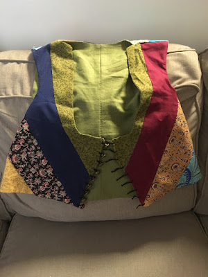 A striped patchwork vest with green lining and green, gold, red, and blue stripes, and gold buttons with black loops, draped on the back of a tan couch.