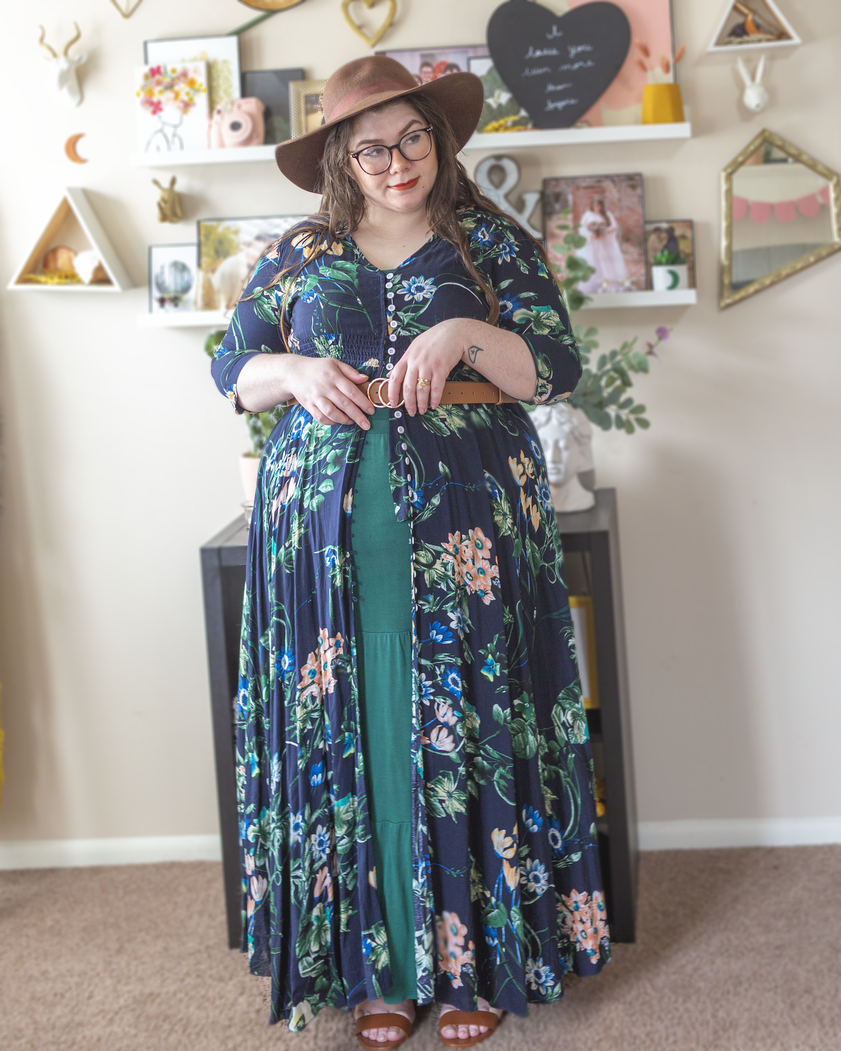 An outfit consisting of a dark brown wide brim floppy hat, 3/4 sleeve navy blue button up maxi dress with a green, yellow, pink and white floral motif layered over a forest green halter maxi dress and belted over with a cognac brown belt and cognac brown heeled ankle strap sandals.