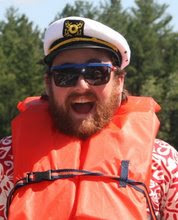 A picture of Mr. Craig, he has a full beard, is making a silly face and is wearing a captain’s hat and a bright orange life-vest.