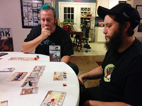 Two players contemplating their next move at Harbour. Both are sitting at a table looking at the components of Harbour, which are spread out before them.