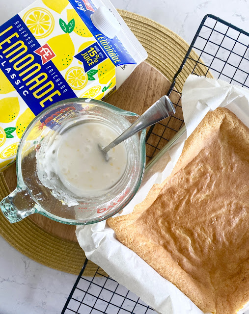 AE Dairy lemonade, a mixing glass of icing and a lemon brownies baked in a baking dish all sitting on a wooden cutting board.