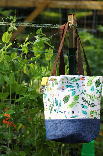 Herb garden patterned project bag with a denim boxed bottom and brown cotton webbing dual shoulder straps. Two orange heart snap buttons on top for closure. Yellow loop on top edge with stitch markers on rectangular ring. Set on the pole holding up strings for thriving pea plants.