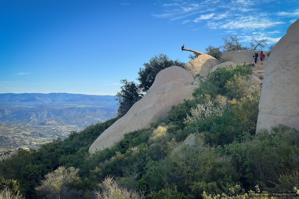 Someone sitting on a distant Potato Chip Rock close to the summit of Mount Woodson with green busy chapparal in the foreground, and jagged mountains in the background.