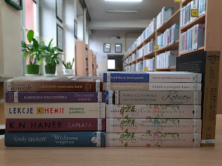 Na fotografii znajduje się jedenaście książek ułożonych na biurku poziomo jedna na drugiej w dwóch kolumnach. W tle regały biblioteczne.