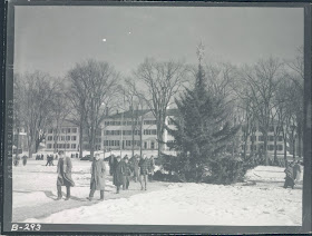 Black and white photo of college students walking to class on the green in the snow with the christmas tree in the background