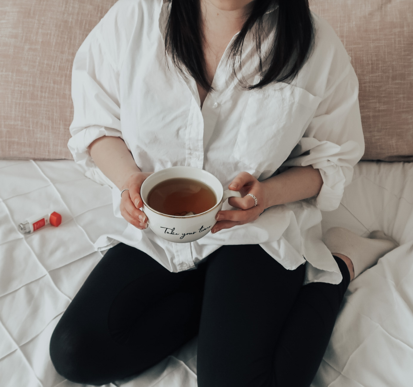 A woman holding a white mug sat on a bed next to an inhaler.