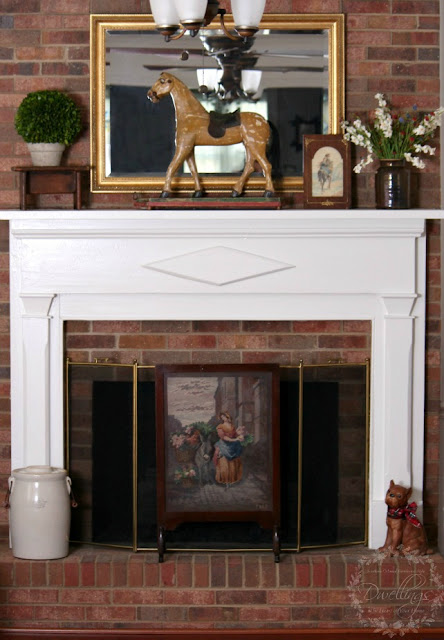 On the fireplace is an antique horse pull toy, old crock filled with flowers, a watercolor horse painting and a needlework fireplace screen with a lady gathering flowers and placing in them in a basket on the back of a donkey.