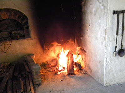 Large 18th-century cooking fireplace with wood fire burning. Beehive oven in wall and metal cooking utensils are also seen.