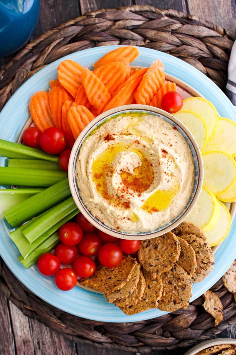 A bowl of hummus on a blue plate surrounded by crackers and chopped vegetables including cherry tomatoes, carrots, celery, and yellow squash on a dark wood table.