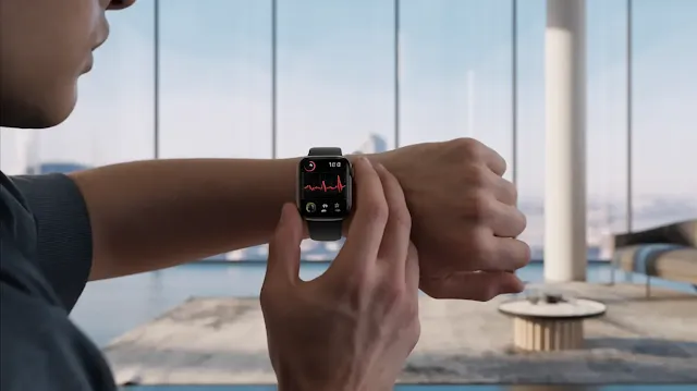 A cinematic shot of a man wearing the Apple Watch X on his wrist. The watch is on his wrist, and it is displaying a heart rate monitor graph. The man is standing in a modern, minimalist room with a few pieces of furniture. The room has large windows with a view of a city.