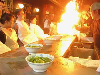 3 people sitting behind a counter with white bibs on. There are white bowls in front of each person with ramen noodles and green onions. Behind the counter is a man in a short chef's hat holding a wok that is tilted toward the people sitting behind the counter. The wok is filled with oil and on fire. A large fireball sits above the white bowl between the chef and the patrons.