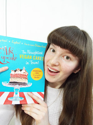 Ellie smiling brightly, holding up a Ms Cupcake vegan baking book, pointing to the cake on the cover with practically a sparkle in her eyes