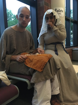 A seated, nearly bald young white man in a grey tunic and white loose pants, with an orange napkin and cheese sticks with plastic wrappers on his lap. A young Indian woman is seated next to him, in a lighter grey dress with a blue sash tied at her side and a sheer white veil. They've both been caught eating; he's looking down away from the camera, and she's covering her mouth with a hand.