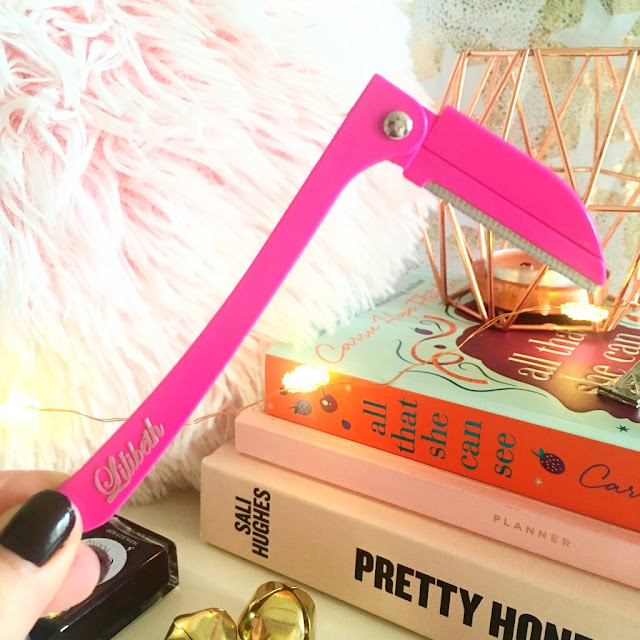 Opened brow shaper held up in front of pink fluffy pillow, stack of books and copper candle holder