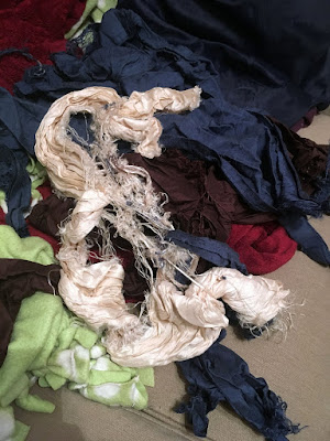 A pile of crumpled and twisted cream, deep blue, and brown silk fabric on a white-spotted green fleece blanket and a red plush blanket. Long frayed threads from each of the silk pieces, but especially the cream, are tangled throughout the whole mess.