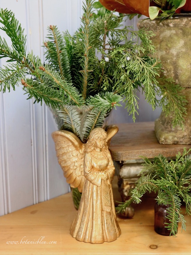 An angel flower vase holding fresh evergreens will be replaced with a simpler mercury vase to transition from Christmas to Winter decor