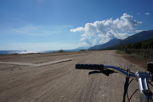 Left-half of a bike handlebar amidst a dry dirt road and a background of blue skies and a mountain