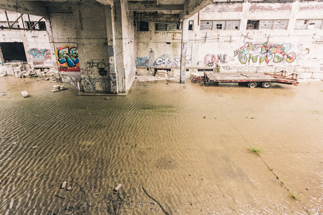 An indoor expanse of an old factory flooded with shallow water, reflecting a worn interior punctuated by structural columns. Graffiti art and tags cover the walls, providing a stark contrast to the industrial decay. A lone flatbed trailer sits marooned on the concrete floor, adding to the scene's post-industrial desolation.