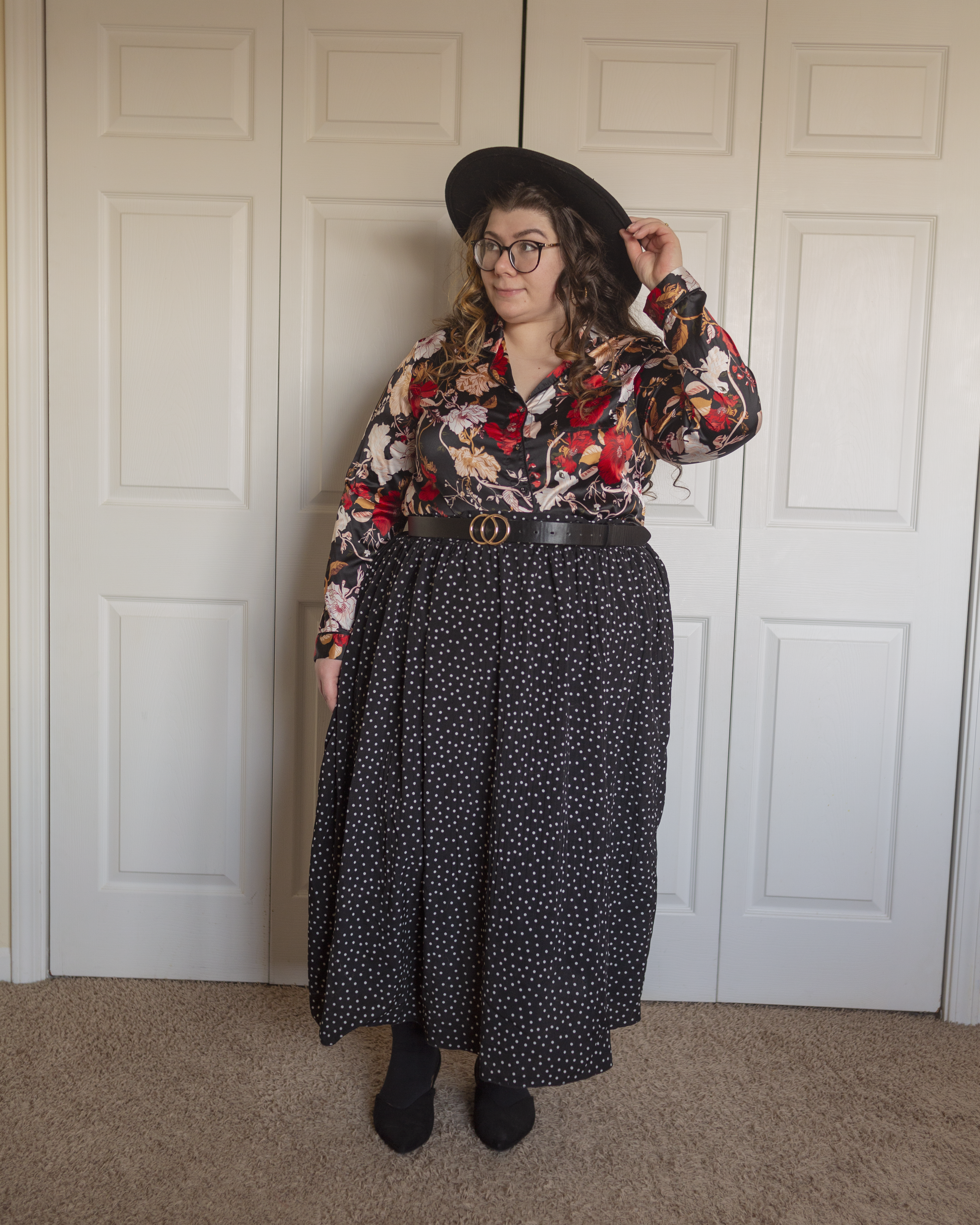 An outfit consisting of a wide brim black porkpie hat, a black, red, pastel pink, mustard yellow and champagne floral notched collar pajama style blouse tucked into a white on black swiss dot midi skirt and black sling back flats.
