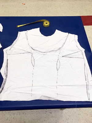 A slightly crumpled white muslin with sharpie marking the seam lines, mismatched bust darts, and tentative neckline, laid flat on a blue table with a yellow tape measure spooled above the neck opening.