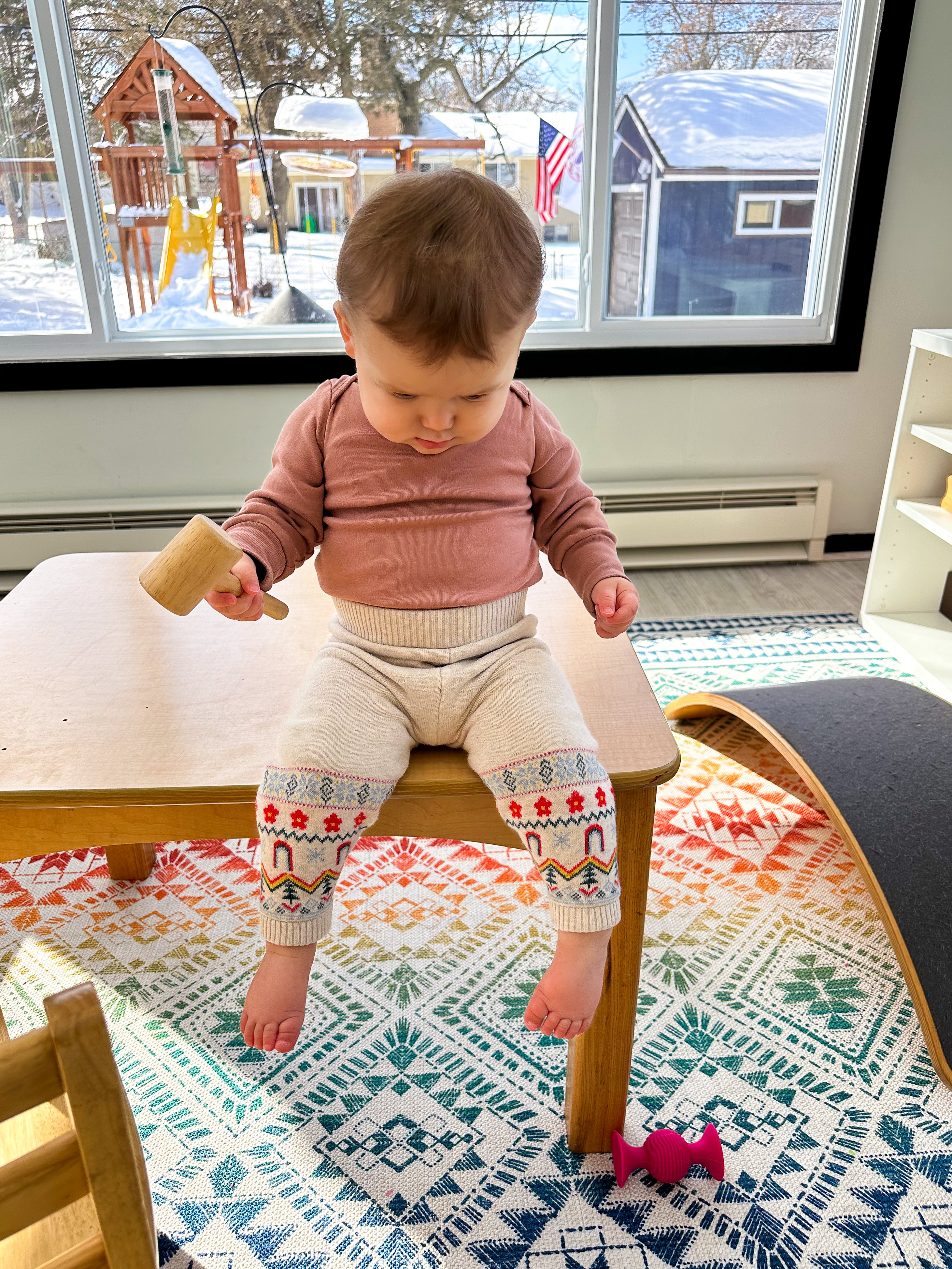 Montessori baby sits on top of table while holding a toy and looking toward her feet and the floor.