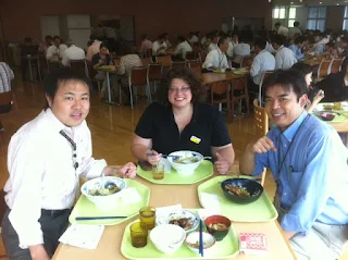 3 people sitting at a wodden table with bowls of food in front of them. The person on the right is a man with black hair, smiling, and wearing a white long sleeve dress shirt and dark pants with a black lanyard keychain. Next to him seated at the center of the photo is a woman with dark brown curly harir and glasses wearing a short sleeve black polo style shirt. Next to the woman on the right side of the photo is a man with black hair wearing a light blue dress shirt.