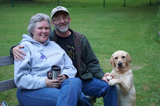 A woman and a man sit on a bench outside. The man has his arm around the woman. A yellow Labrador retriever sits and rests her front paws on the man's knee.