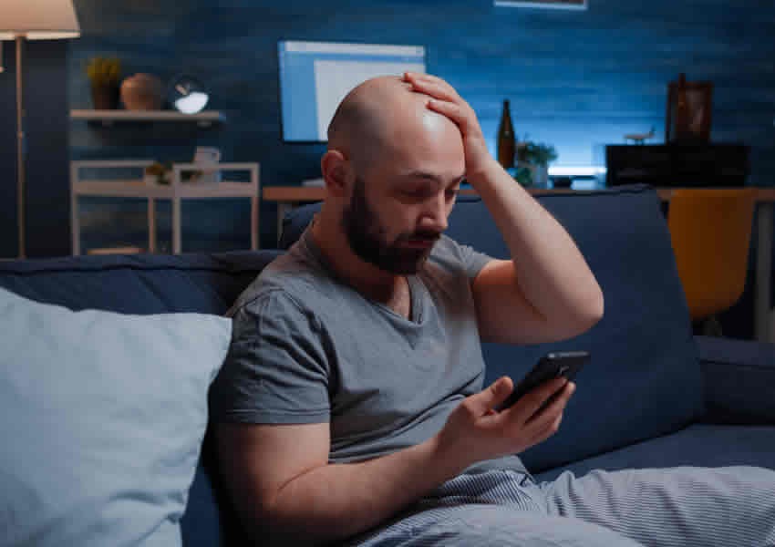 Homem sentado no sofá com rosto de preocupado com uma das mãos na cabeça e outra segurando um Smartphone.