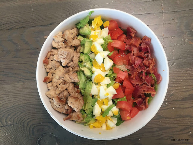 White bowl on wooden table with ground chicken, avocado, hard boiled eggs, tomatoes, bacon and romaine