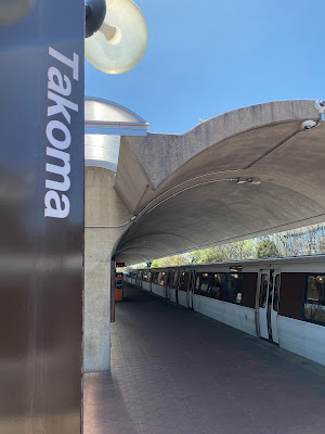 Photo of a train at the platform, which has an arched cover. In the foreground is a metal pillar that says Takoma.