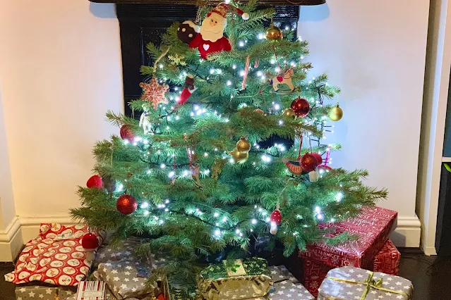 A real Christmas tree in front of a fire place surrounded by presents