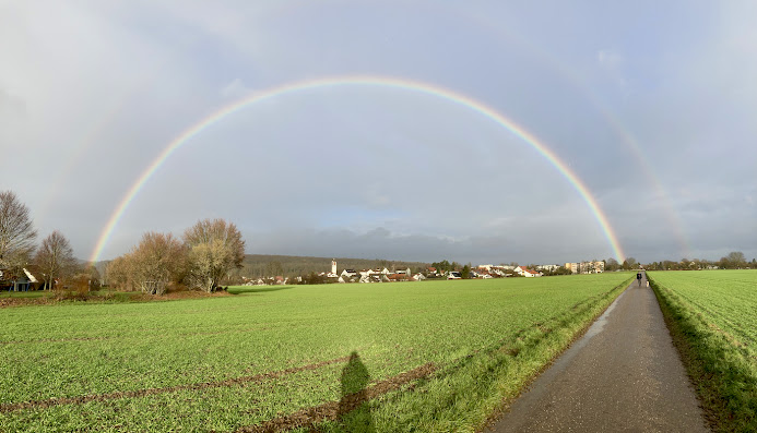 Regenbogen, Acker, Feldweg, Mann mit Hund in der Ferne, noch weiter weg liegt ein Dorf