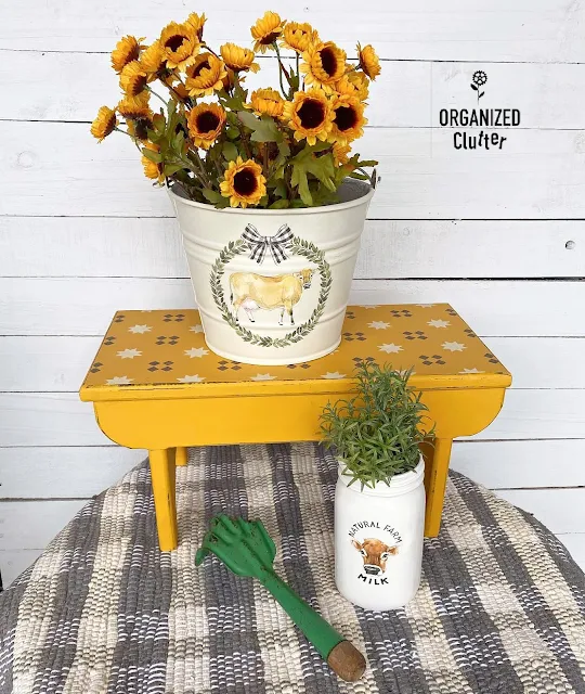 Photo of a painted and stenciled farmhouse bench, with upcycled bucket & jar.