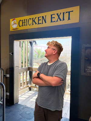 A light-skinned man with gray-blond hair wearing a light blue t-shirt and dark shorts is standing in front of a doorway with a rollercoaster in the background. Above the doorway is a yellow sign that says "Chicken Exit."