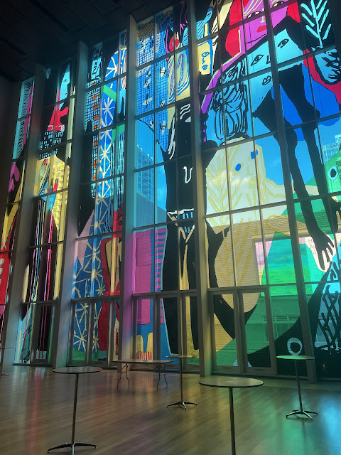 A wall in the museum that separates the inside from the outside of the building that is at least two stories tall (maybe 3) and is one big stained glass picture. The picture is some abstract drawing of city buildings, faces, black bodies, snowflake type designs.