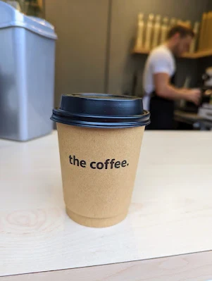 Takeaway coffee cup with lid. Cup says "The Coffee" on the front. Blurred barista in the back right