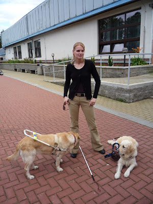 Linda stojí před hotelem. U nohou leží zlatý retrívr a vedle stojí světlý labrador v postroji