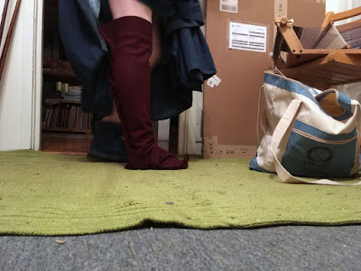 A photo from floor level of a person's leg from the knee down, wearing a garnet-red hose with small wrinkles at the knee, and large intersecting bubbles and wrinkles at the ankle and over the instep, with a tightly curled toe. The person's other foot is half-hidden behind, in a blue quilted house slipper, and they're holding their long denim skirt up to show their leg. A ribbed bright green rug covers most of the floor, and a white and light-blue zipper tote from A Novel Idea slouches to the right of the person.