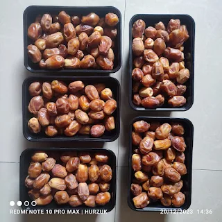 A diverse group of volunteers, including men and women of different ethnicities, unload boxes of dates from a van in a busy Ramadan marketplace. Festive decorations and smiling faces create a cheerful atmosphere.