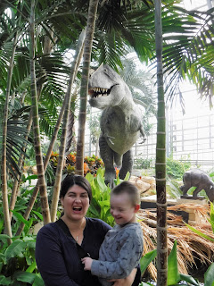 a woman roars while a boy laughs, and a tyrannosaur statue and large tropical plants are visible in the back