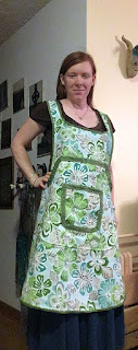 The first of four images in a row, showing a white, redheaded woman at the corner of a hallway, wearing a green, turquoise, and grey bibbed apron bound in light lime-green, with a large near-trapezoidal pocket and a small rounded-square pocket center front, also bound in light lime-green, over a brown t-shirt and long denim skirt. Her left hand is propped on her hip, just at the edge of the apron, and she's grinning.